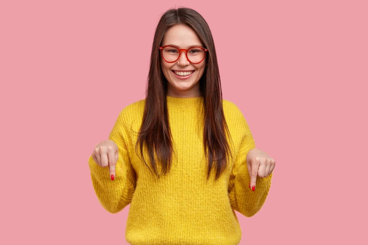 Young woman with long dark hair wearing a yellow jumper with a positive expression and is pointing down with both index fingers.