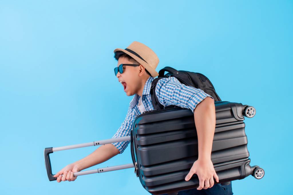 Casually-dressed young man wearing a straw hat and sunglasses carrying a suitcase under his arm with an excited expression and open mouth.