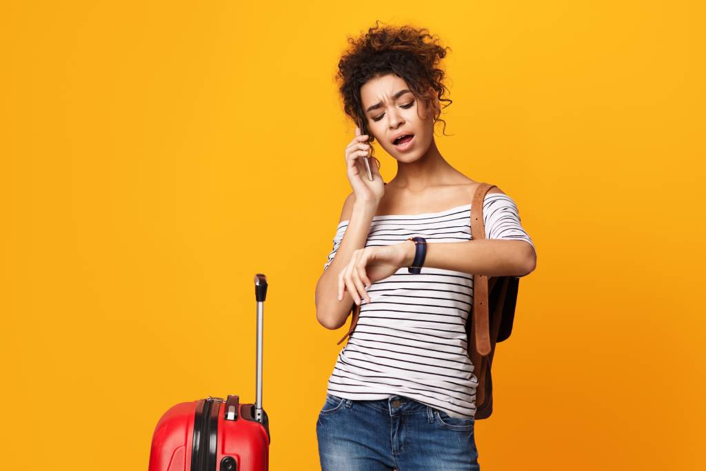 Young woman with short, dark curly hair wearing jeans and a striped T-shirt with a frustrated expression and looking at her watch, with a suitcase by her side.