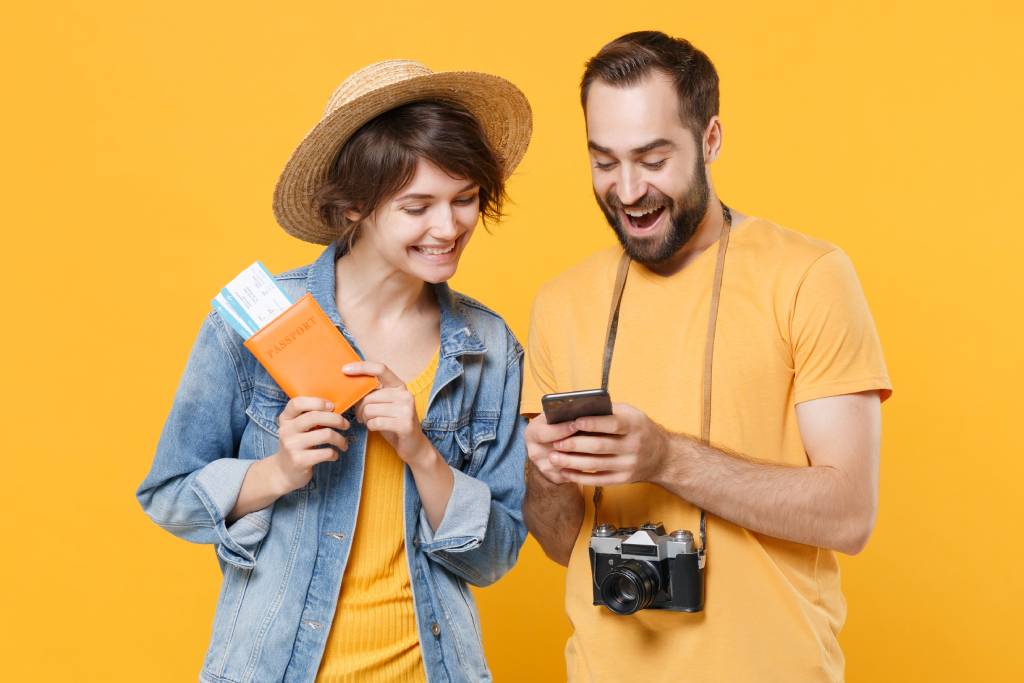 Woman wearing a denim shirt and a straw hat holding travel tickets in her hands beside man with short dark hair and a beard with camera around his neck, both smiling at a mobile phone in the man’s hands.