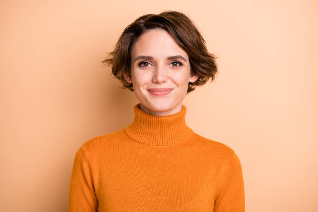 Young woman with short dark hair wearing an orange turtleneck jumper and smiling straight ahead