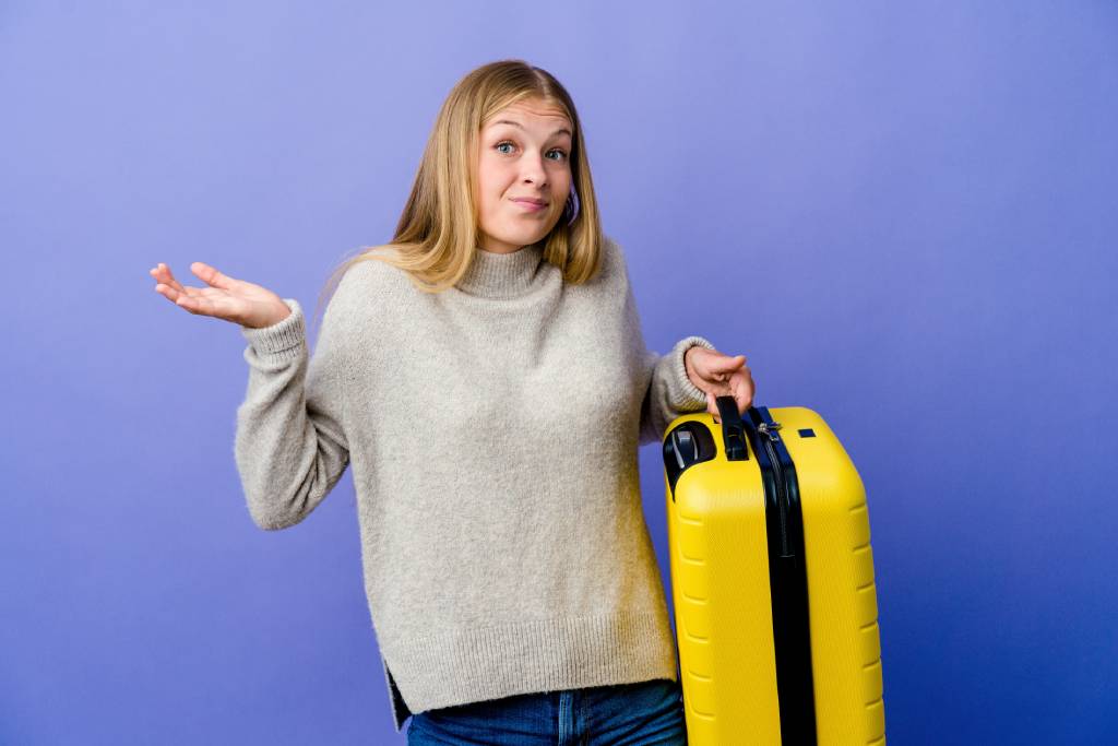 Young woman with long fair hair wearing a grey jumper holding a yellow suitcase in her left hand and holding her right hand up, shrugging her shoulders with a perplexed expression.