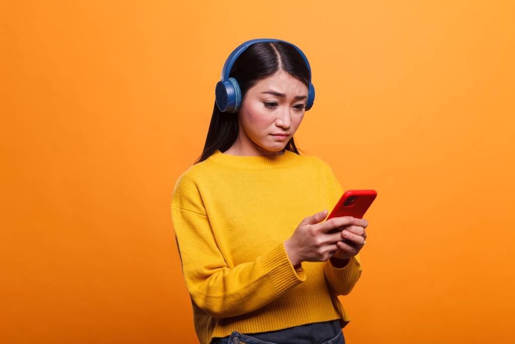 Young woman with long dark hair wearing a yellow jumper wearing blue headphones looking at a mobile phone with a puzzled expression.