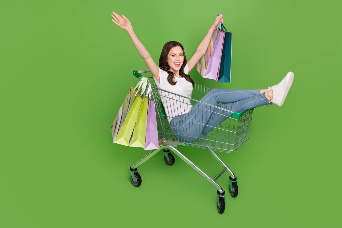 Fun-loving young woman wearing jeans and a white T-shirt holding shopping bags, seated in a shopping trolley