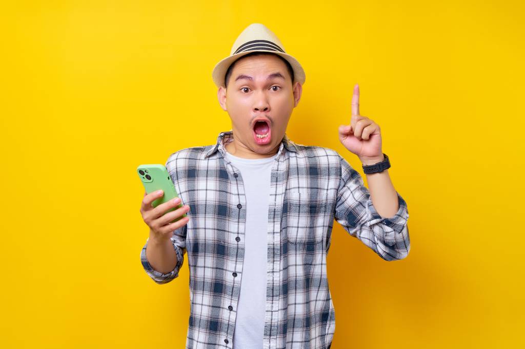 Young man wearing a white T-shirt and checked over-shirt and a straw hat holding a mobile phone in one hand and pointing the index finger of his other hand upwards. He has a shocked expression on his face and his mouth is wide-open.
