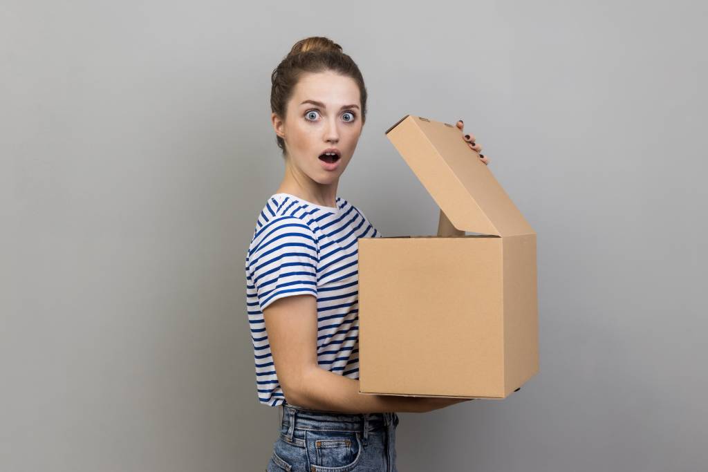 Side-view of a young woman with dark hair in a bun wearing a striped T-shirt holding a cardboard box with a surprised expression.