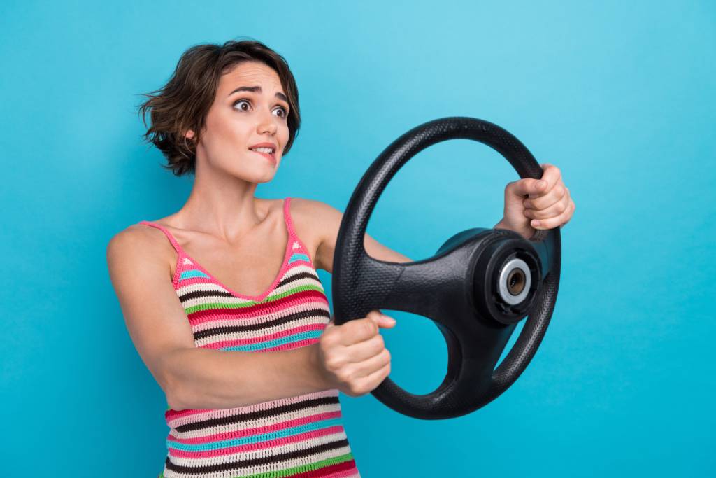 Young woman with short dark hair wearing a striped tank top holding a car steering wheel out in front of her with a scared expression and biting her lip.
