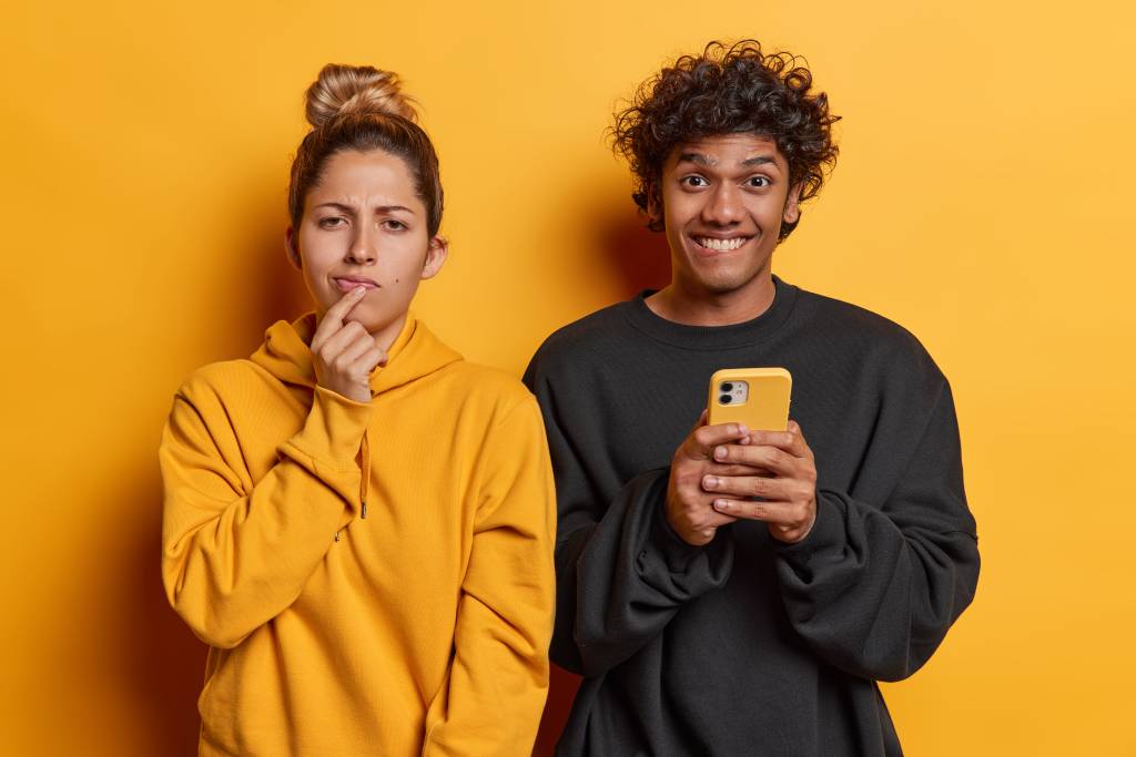 Woman with dark hair in a bun wearing a yellow sweatshirt with a puzzled expression standing next to a man with curly dark hair wearing a black sweatshirt, holding a mobile phone and smiling.