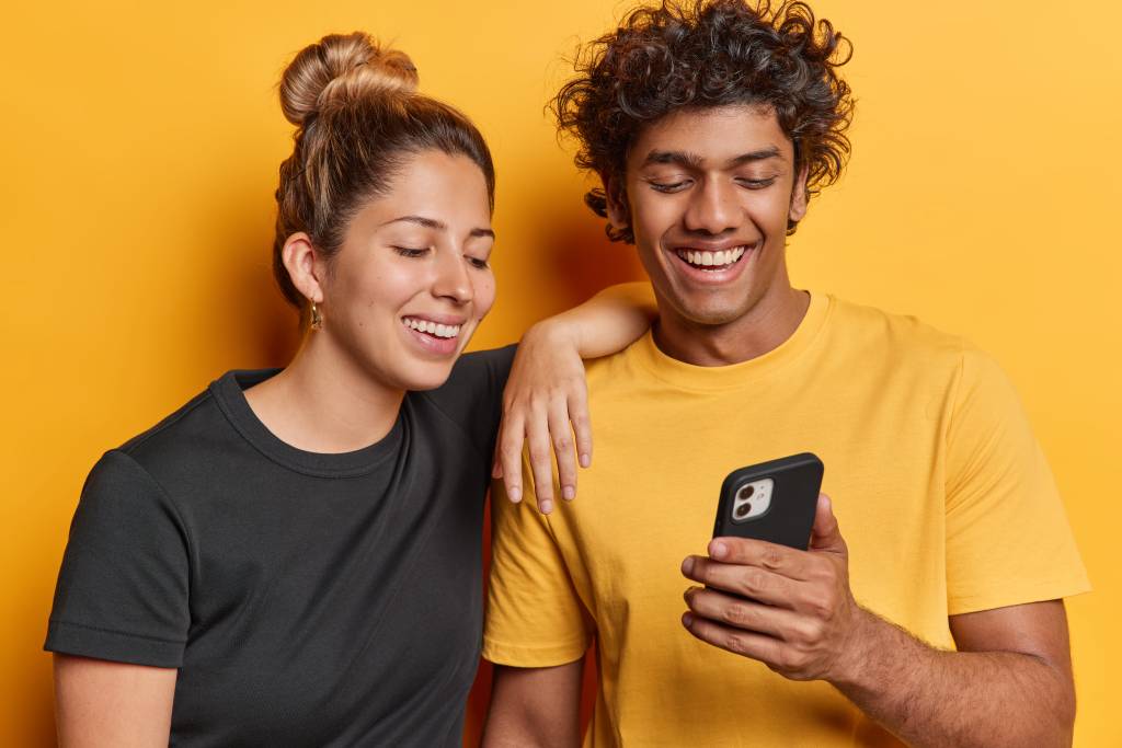 Woman with dark hair in a bun wearing a black T-shirt standing next to a man with curly dark hair wearing a yellow T-shirt sweatshirt, both smiling at a mobile phone in the man’s hands.